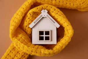 A small wooden house model is wrapped in a cozy, bright yellow knitted scarf, symbolizing warmth and home comfort, against a soft beige background.