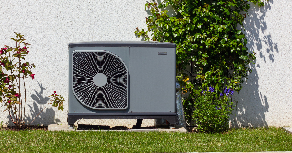 A gray outdoor air conditioning unit, ideal for heat pumps in dry climates, sits on a concrete pad by a white wall, surrounded by green grass, flowering plants, and leafy bushes.