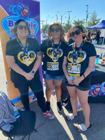 Three women wearing matching black shirts, sunglasses, and race bibs stand outdoors at an event. Behind them is a Bumble Bee promotional banner and a table with event materials. It is sunny and bright.