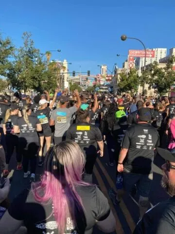 A crowd of people, many wearing black shirts, participate in an outdoor race or walkathon on a sunny day, walking down a city street lined with trees and buildings.