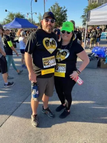 A man and a woman wearing matching black race shirts and numbered bibs pose together at an outdoor race event, surrounded by other participants and tents under a sunny sky.