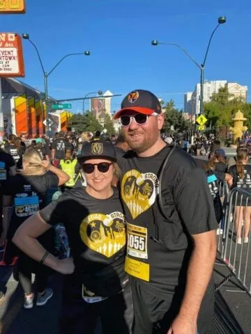 Two people wearing matching black shirts and hats with yellow designs and race bibs, smiling at an outdoor event with a crowd of runners on a sunny day.