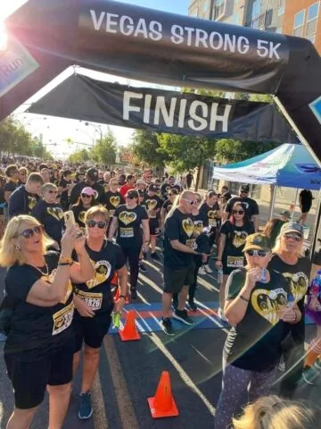 A large group of people in matching black shirts with gold heart designs gather at the finish line of the "Vegas Strong 5K" race, smiling and taking photos under a sunny sky.