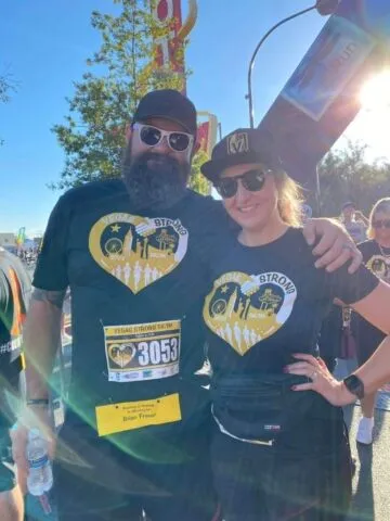 Two people in matching black shirts and hats with yellow designs stand outside at a race event, smiling at the camera. One wears sunglasses and a beard, the other sports a fanny pack and bib number 3053.