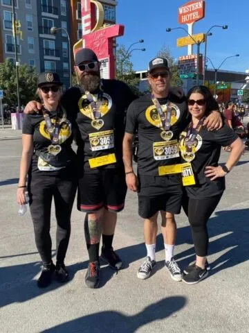Four people in matching black shirts and medals stand together smiling after a race, with race numbers on their shirts. Urban street scene in the background with signs and buildings visible.