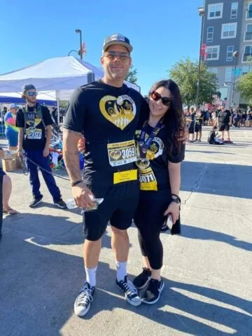 A man and woman wearing matching black race shirts and bib numbers pose together at an outdoor event, smiling at the camera. Other participants and tents are visible in the background under a clear sky.