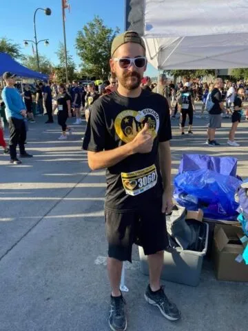 A man wearing sunglasses, a cap, and a black T-shirt with a race bib stands outdoors at an event, giving a thumbs-up. Other people and tents are visible in the background on a sunny day.