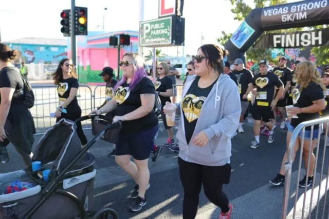 A group of people, some wearing matching shirts, walk past the finish line at a race event. One person pushes a stroller, and others carry drinks. The background shows colorful buildings and event signage.