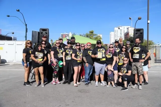 A group of people in matching black shirts with a yellow logo pose together outdoors on a sunny day, some holding a certificate, with speakers and buildings visible in the background.