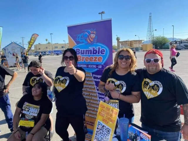 A group of smiling people in matching black shirts pose outdoors in front of a "Bumble Breeze" Plumbing, Heating, and Air sign at an event, with booths and banners visible in the background.