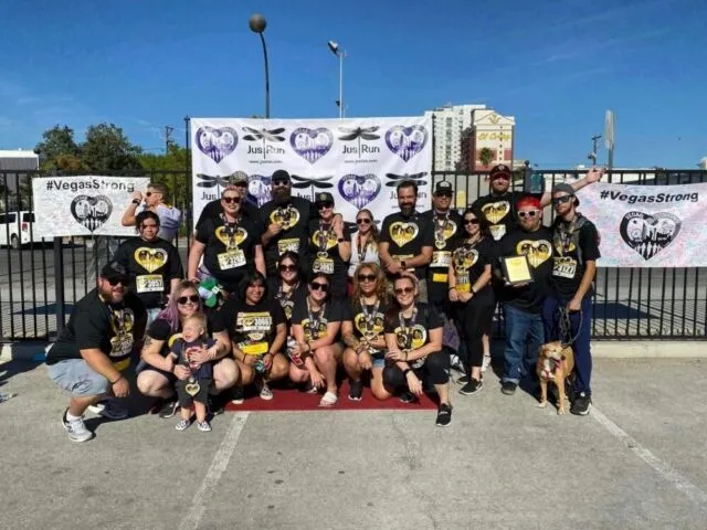 A large group of people, many wearing matching black "Vegas Strong" shirts, pose together outdoors in front of a banner at an event. Some hold medals and awards, and a dog in sunglasses is in the front row.