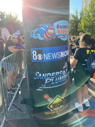 People gather around a large inflatable pillar with logos for Bumble Breeze Plumbing, 8 News Now, Sanders Plumbing, and Viome Masters of Bad Decisions. Sunlight creates a lens flare across the image.