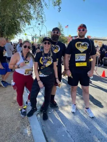 Four people wearing Batman-themed shirts and race bibs pose together at an outdoor event, possibly a charity run, with other participants and trees visible in the background.