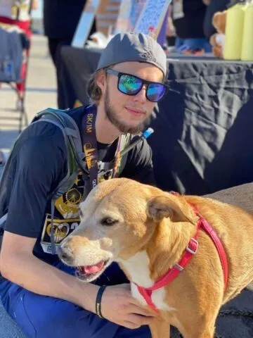 A man wearing sunglasses and a backward cap kneels beside a brown dog with a red harness. They are outdoors at an event with tables and items in the background.