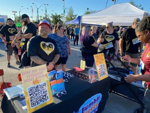 People gather around a booth at an outdoor event. Two signs with QR codes and flyers are on the table. Attendees, some in matching shirts, stand nearby under tents on a sunny day.