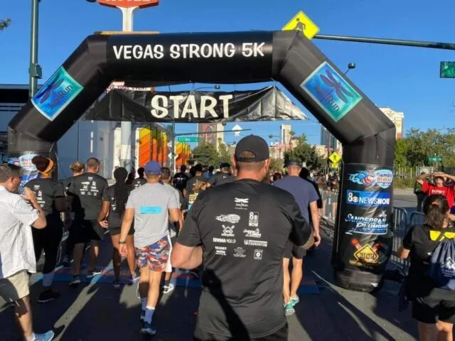 A group of runners in athletic gear walk under a large black inflatable arch with "START" and "Vegas Strong 5K" written on it, beginning a race on a sunny day in a city setting.