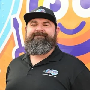 A man with a full beard and mustache wears a black cap and black polo shirt with a "Plumbing Heating Air" logo, standing in front of a colorful background featuring a smiling cartoon face.