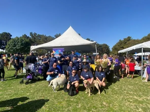 A group of people in matching shirts poses with two dogs in front of a white tent at an outdoor event on a sunny day, surrounded by other attendees and trees.