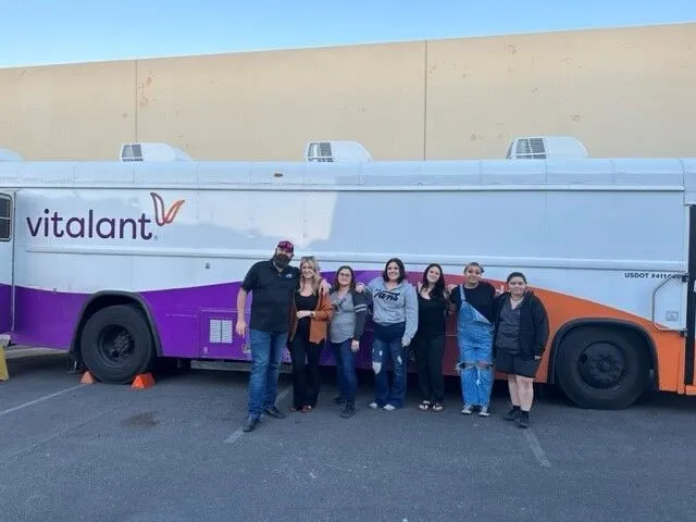 A group of seven people stand smiling in front of a white and purple Vitalant blood donation bus parked outside on a sunny day.