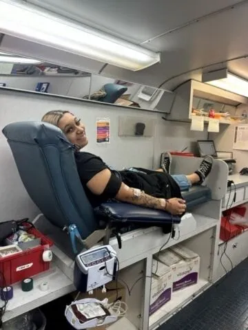A person is lying on a reclined chair in a clinic, donating blood. They are smiling at the camera, with a blood pressure cuff and tubes attached to their arm. Medical supplies are visible nearby.
