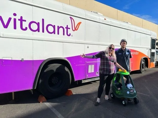 Two smiling adults stand beside a large Vitalant blood donation bus. One person has a hand on a green toy car. The scene is outdoors on sunny day, with shadows on the pavement.