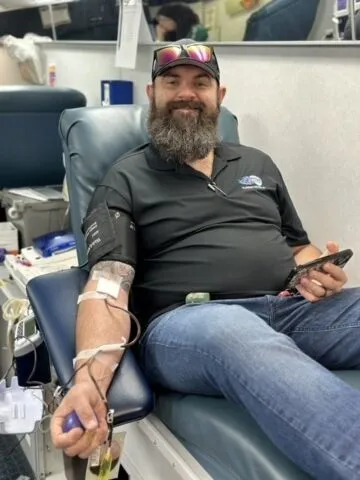 A bearded man wearing a black shirt and jeans sits in a chair, smiling while donating blood. He holds a phone in one hand and has a needle and tubing attached to his other arm.