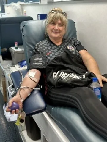 A woman sits in a medical chair donating blood, smiling at the camera. She holds a stress ball in one hand, with medical tubes attached to her arm. A water bottle and personal items are on the chair beside her.