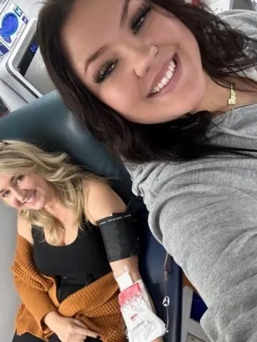 Two women smile for a selfie in a medical setting. One is seated in a chair with a blood pressure cuff and a bandaged arm, possibly after donating blood or receiving treatment. Both appear happy and relaxed.