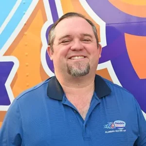 A man with short brown hair and a goatee smiles while wearing a blue "Bumble Breeze Plumbing Heating Air" shirt, standing in front of a colorful orange and purple background.