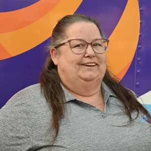 A smiling woman with long brown hair and glasses wears a gray collared shirt, standing in front of a colorful background with purple and orange curved shapes.