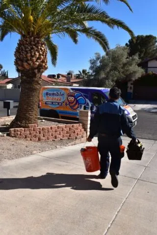 A worker carrying a red bucket and black toolbox walks toward a Bumble Bee-branded pest control van parked on a residential street, with palm trees and houses in the background.