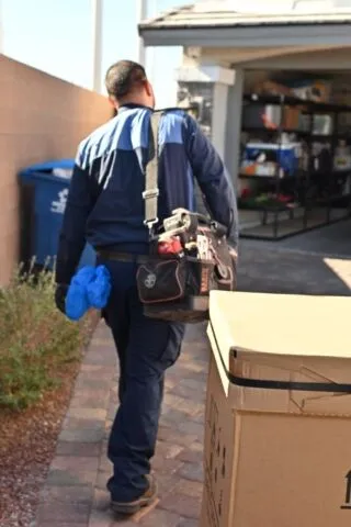 A person in a blue uniform carrying a tool bag walks along a pathway toward a garage, passing a large cardboard box and a blue recycling bin.