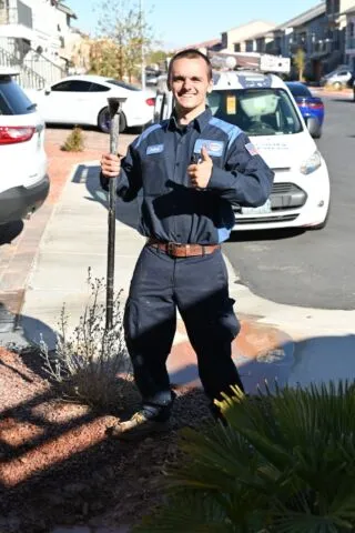 A smiling plumber in uniform holds a large wrench and gives a thumbs-up while standing outside on a sunny residential street, with service vehicles and houses in the background.