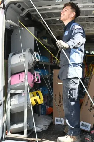 A technician in a work uniform stands inside a van filled with equipment and colored gas tanks, holding long metal rods and smiling while looking up.