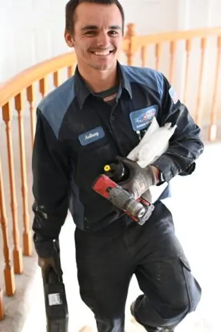 A smiling maintenance worker in a navy uniform carrying tools and supplies stands indoors near a wooden staircase railing.