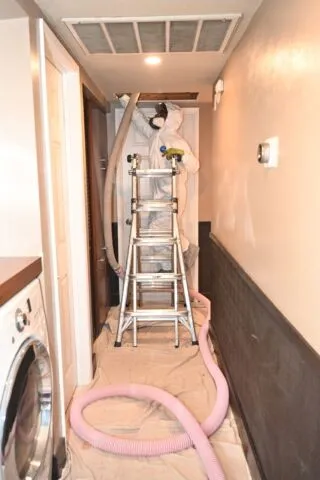 A worker in protective coveralls stands on a stepladder under an open vent, using a hose for cleaning. Pink and white hoses extend onto a covered floor in a narrow hallway near a washing machine.