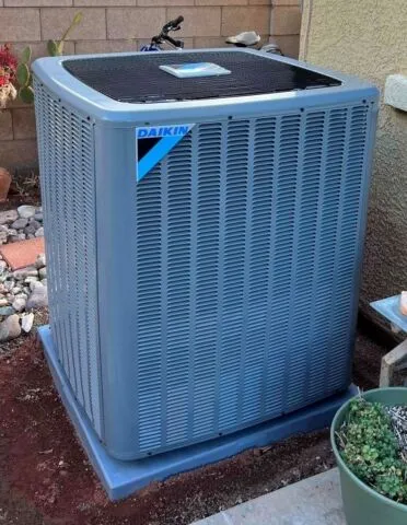 A Daikin outdoor air conditioning unit sits on a concrete pad next to a house, surrounded by gravel, rocks, and plants.
