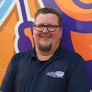 A man with glasses and a beard smiles at the camera while wearing a navy blue polo shirt with a "Bumble" logo. He stands in front of a colorful orange and purple background with abstract designs.