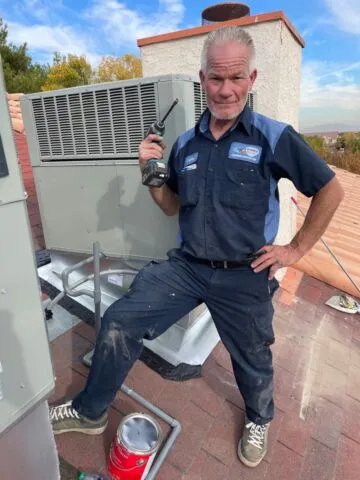A maintenance worker in a navy uniform stands on a rooftop next to an HVAC unit, holding a cordless power drill and looking at the camera, with one foot resting on a red can.