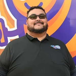A man with short dark hair, a beard, and sunglasses smiles while standing in front of a colorful purple and orange background. He is wearing a black collared shirt with a logo on the chest.