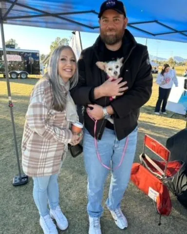 A smiling woman and a man stand together under a tent outdoors. The man holds a small white dog. The woman wears a plaid jacket and the man wears a dark jacket and cap. There are tents and people in the background.