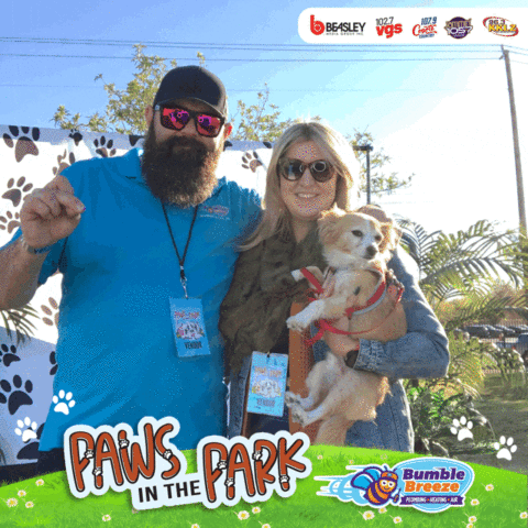 A man and woman smile at an outdoor event, with the woman holding a small tan and white dog. "Paws in the Park" and "Bumble Breeze" logos are at the bottom of the image. Paw print decorations are in the background.