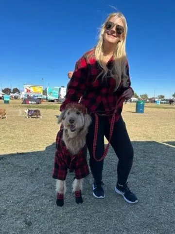 A smiling woman in sunglasses and black leggings stands outdoors holding a dog on a leash. Both the woman and the dog are wearing matching red and black plaid jackets. The sky is clear and blue.