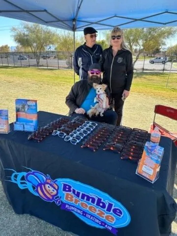 Three people, one holding a small dog, stand behind a table covered with sunglasses and Bumble Breeze promotional materials under a blue canopy at an outdoor event.