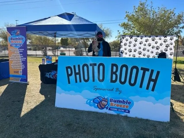 A person holding a small dog stands behind a large "Photo Booth" sign sponsored by Bumble Breeze at an outdoor event with a blue tent and banner in the background.