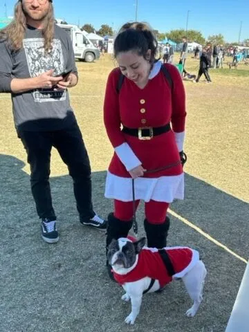 A woman dressed in a Santa outfit stands on grass, smiling down at a small dog also wearing a Santa costume. A man in casual clothes stands nearby, holding a phone. It's a sunny outdoor event.