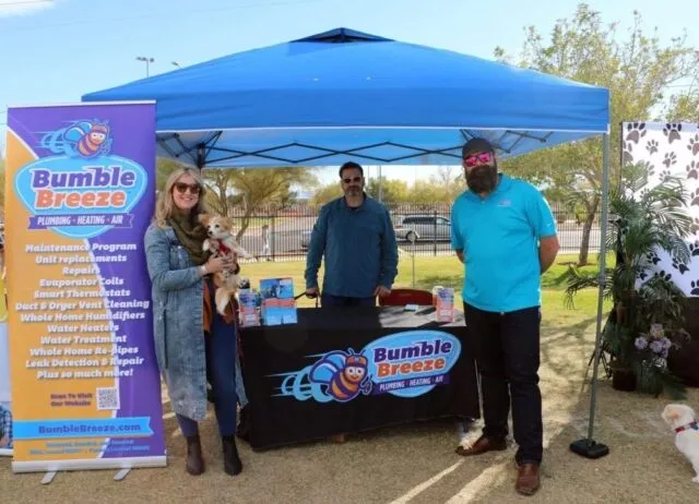 Three people stand under a blue pop-up tent with a "Bumble Breeze Plumbing, Heating, Air" table and banner. One person holds two small dogs. It’s sunny and there are trees and grass in the background.