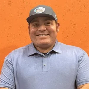 A smiling man wearing a gray baseball cap and a light blue polo shirt stands in front of an orange textured wall.