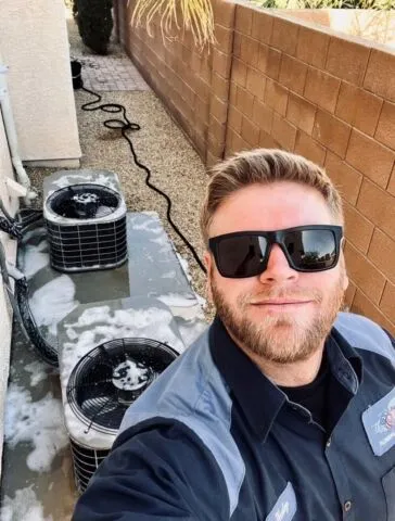 A man wearing sunglasses and a work uniform takes a selfie outside next to two air conditioning units covered in soap and water, indicating they are being cleaned. A brick wall and gravel path are visible in the background.