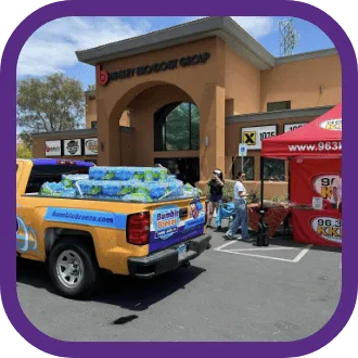 A yellow truck loaded with bottled water is parked outside a building, next to a red tent featuring a radio station logo and an "about us" display. People are gathered nearby on a sunny day.